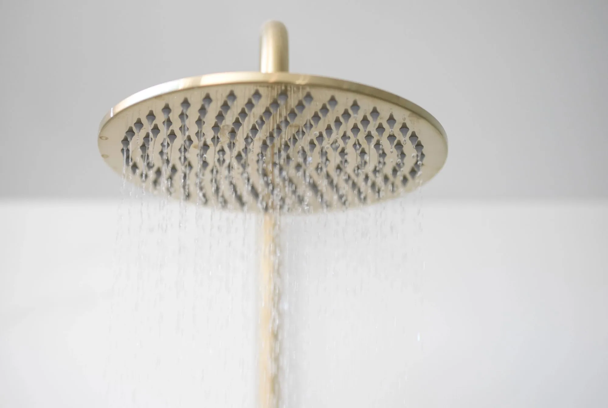 A wide, circular rainfall shower head in a brushed brass finish, mounted against a white wall. 