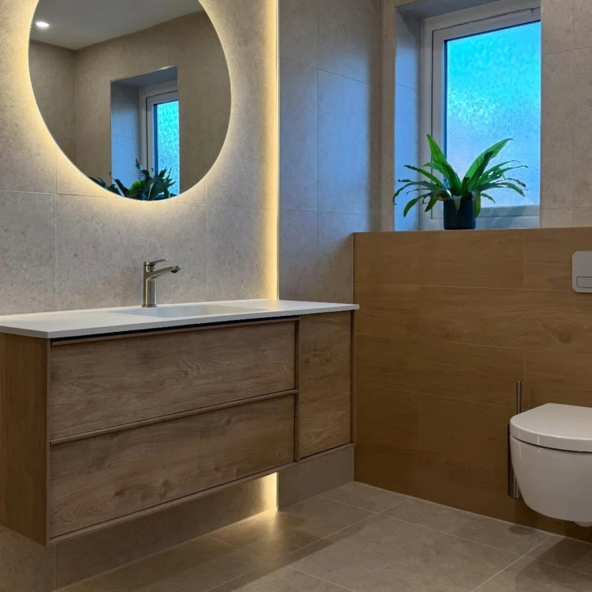 A modern bathroom with stone and wooden finishes featuring a floating vanity beneath a backlit, circular mirror.