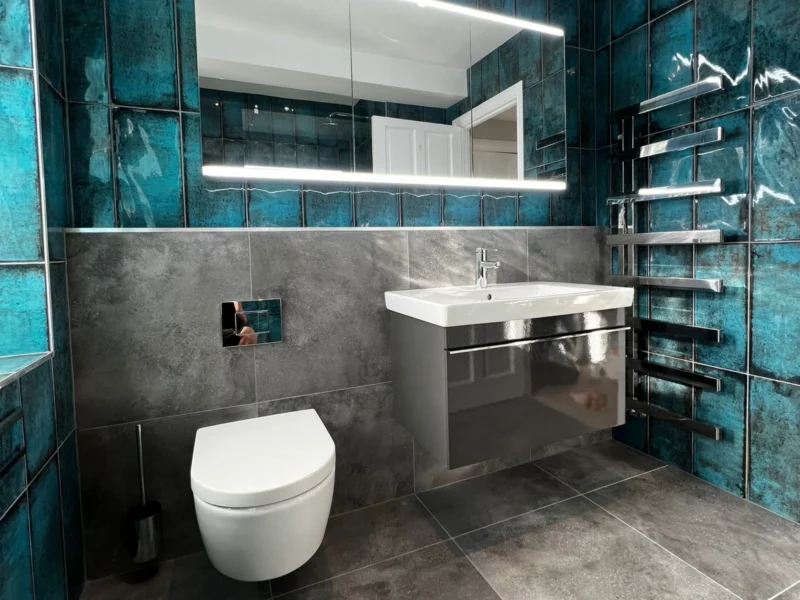 A vibrant modern bathroom featuring glossy teal vertical wall tiles, a grey floating vanity, and a large rectangular LED-backlit mirror.