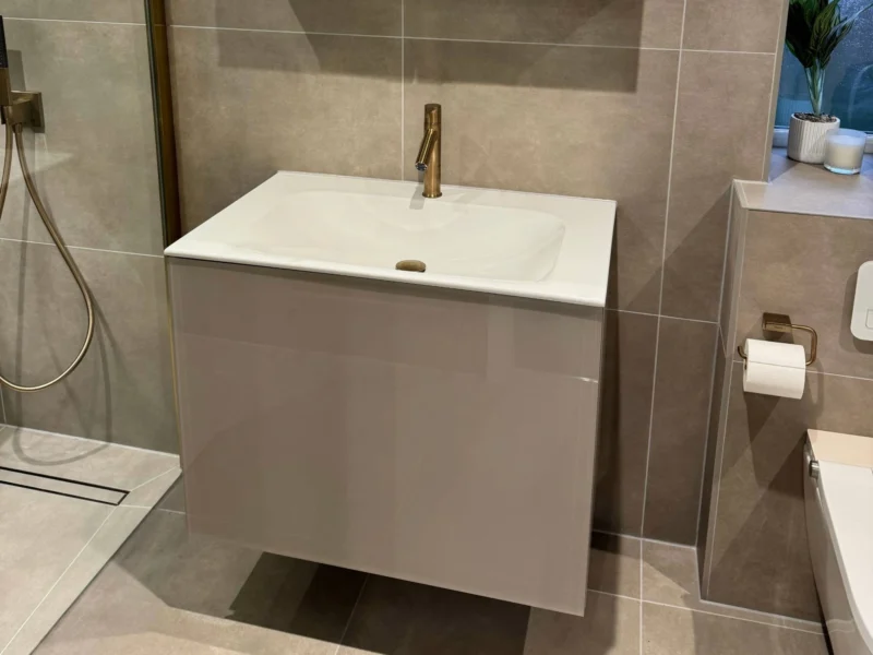 A contemporary bathroom featuring beige wall tiles, a gloss-finish floating vanity with a gold faucet, and a mirrored medicine cabinet with integrated top lighting.
