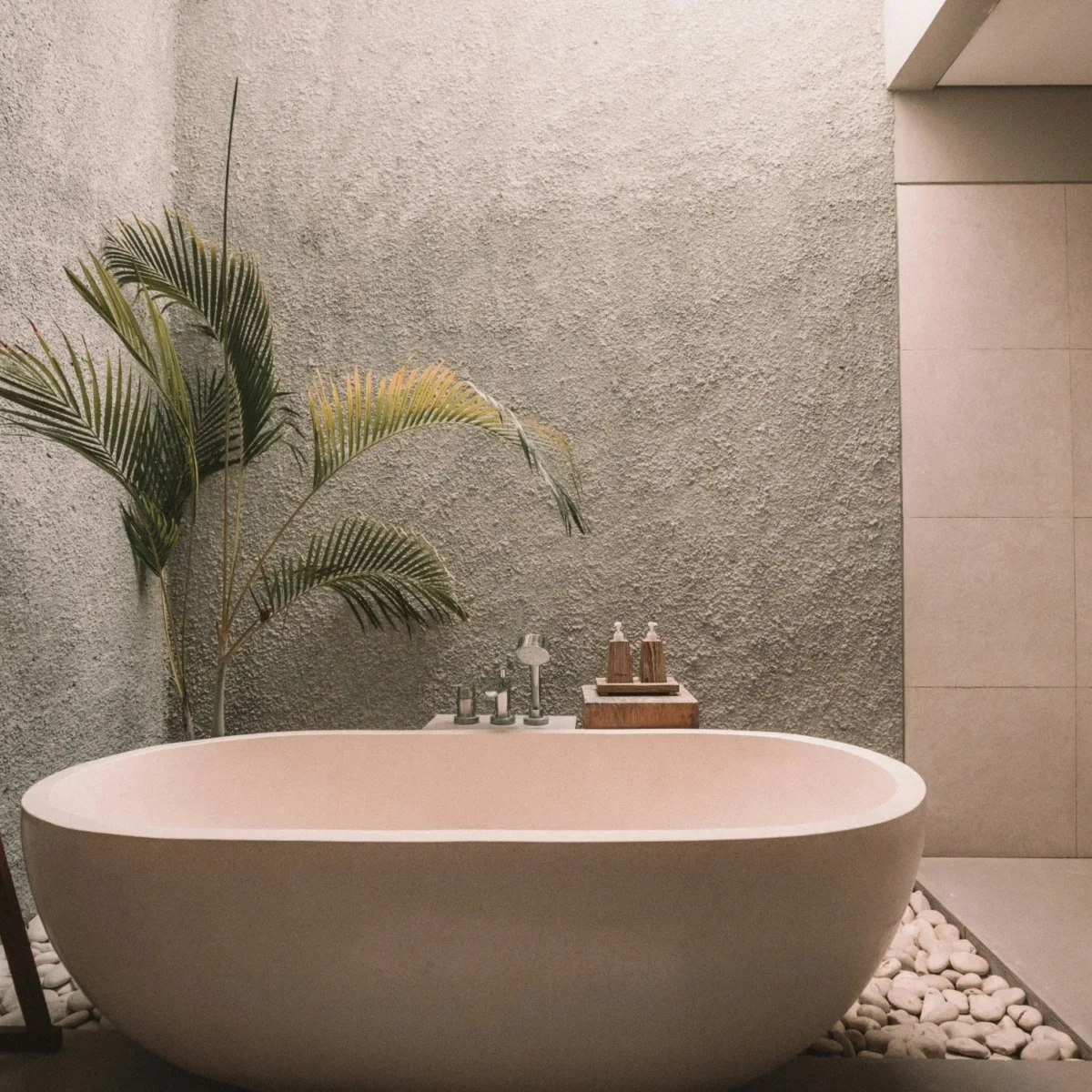 A light pink stone freestanding bathtub in a sparse bathroom with a large plant in the corner.