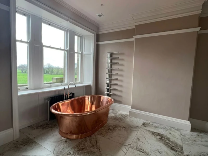 A copper freestanding bathtub sitting in a large bathroom beneath a window with views of the countryside.