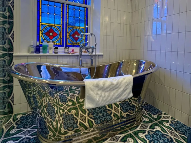 A metallic freestanding bath in a bathroom with colourful wall and floor tiles and stained-glass windows.