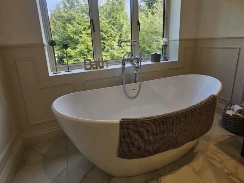 A white freestanding bath in a minimalist bathroom with a window showing a view of green treetops.