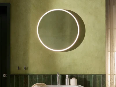 A circular LED vanity mirror upon a mossy green wall above a bathroom sink.