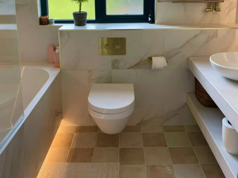 A modern bathroom with white stone effect walls, a wall-mounted toilet, brushed brass towel heater, and neutral square floor tiles. 
