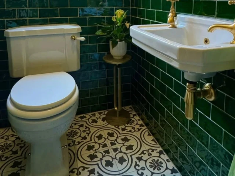 A downstairs bathroom in a maximalist style with walls that are half green tiled and half wallpapered, as well as a stain glass window. 