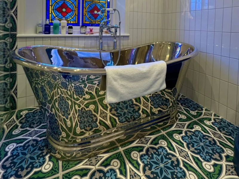 A maximalist bathroom with stained glass windows, a freestanding chrome bath tub, and decorative green, blue, and white tiles. 