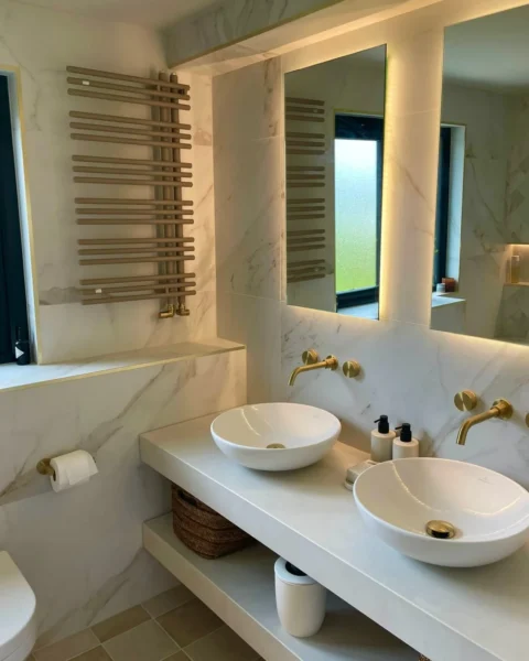 Luxury bathroom with white and gold marble-effect tiles, twin countertop basins, brushed gold wall-mounted taps, backlit mirrors and a matching gold-accented heated towel rail.