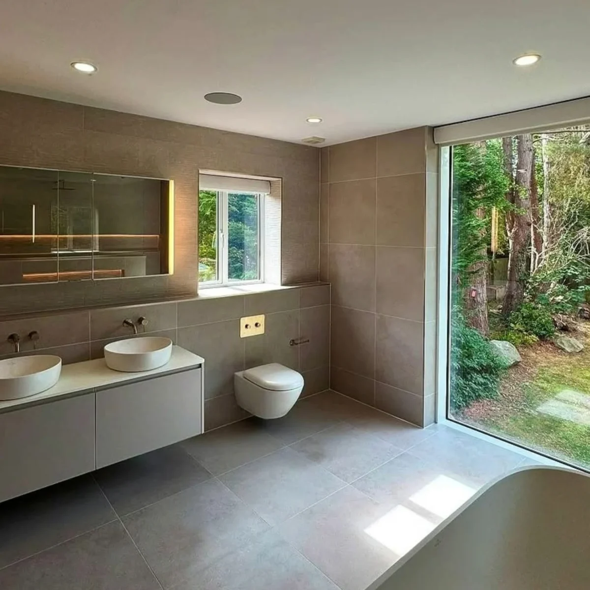 Japandi style walk-in shower with a frameless glass screen, large-format warm stone-effect tiles, an overhead rain shower with handheld attachment, and a softly lit recessed niche.