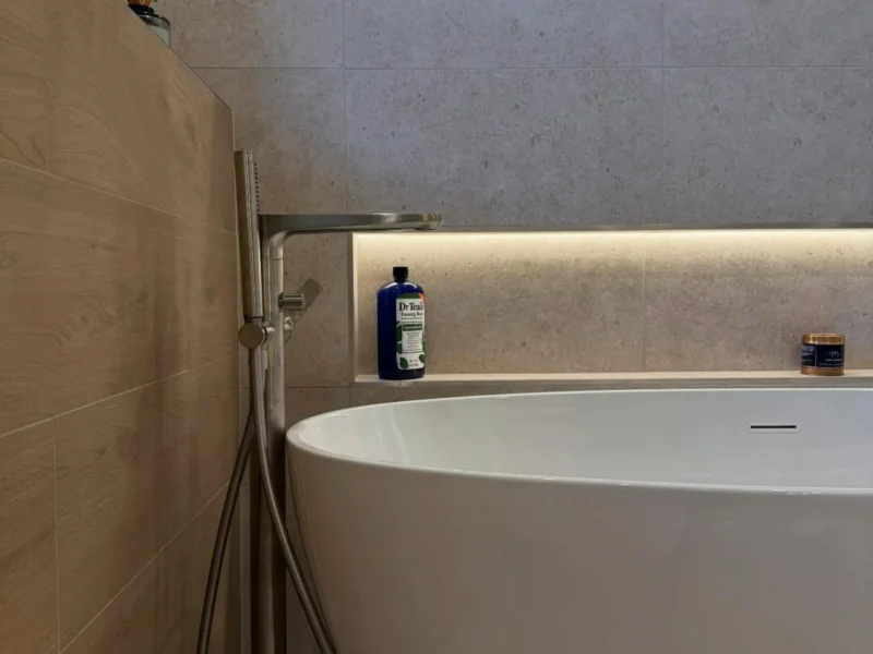 Japandi-inspired bathroom detail of a sculpted white freestanding bath beside a brushed nickel floor-standing tap, with glowing recessed shelves, wood-effect tiles and a trailing green plant.