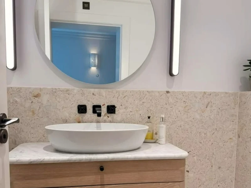 Japandi bathroom vanity in light oak with a white marble top and oval countertop basin, framed by a round mirror, slim black wall lights and speckled terrazzo-effect tiles. 