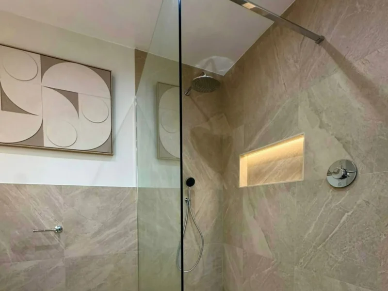 Japandi style walk-in shower with a frameless glass screen, large-format warm stone-effect tiles, an overhead rain shower with handheld attachment, and a softly lit recessed niche. 